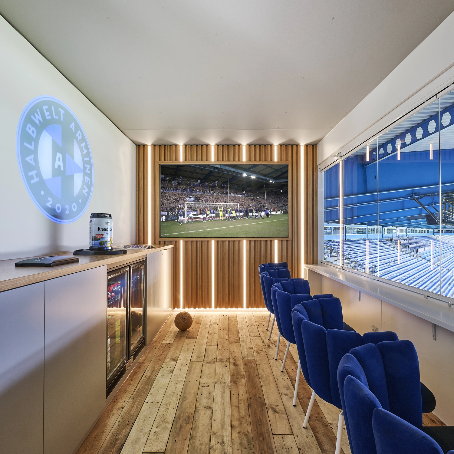 Heinrich Buchholz – Holzbearbeitung – Luxuriöse VIP-Lounge im Stadion mit rustikalem Dielenboden, blauen Samt-Barhockern mit Blick auf das Spielfeld und einem integrierten Bildschirm vor einer modernen Akzentwand aus Holzlamellen mit vertikalen LED-Lichtleisten.