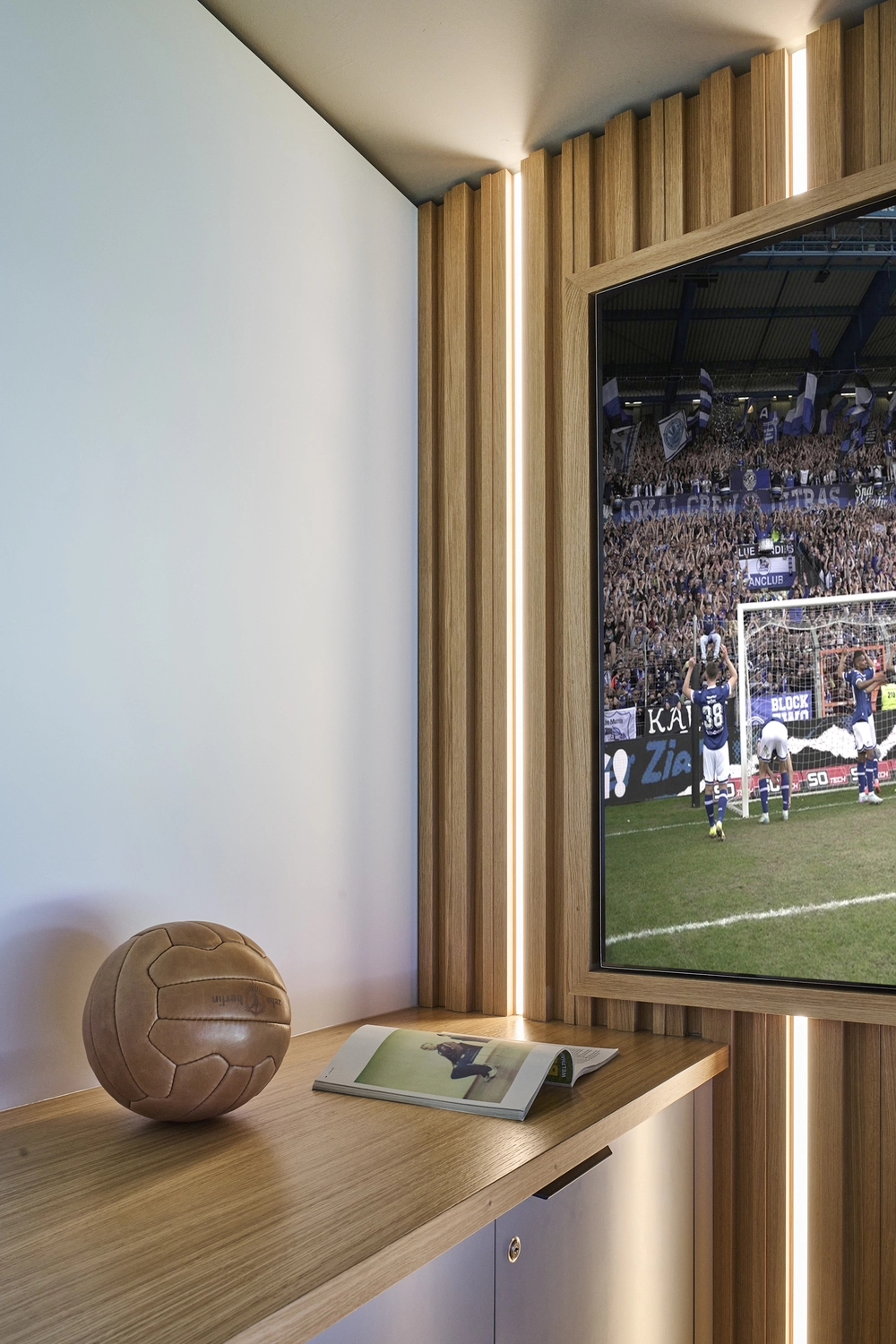 Heinrich Buchholz – Holzbearbeitung – Maßgefertigte TV-Wand mit vertikalen Lamellen aus weißer Eiche und integrierter LED-Beleuchtung, einer Arbeitsplatte aus furniertem Holz mit Kantenanleimung und einem flächenbündig eingelassenen Fernsehrahmen.