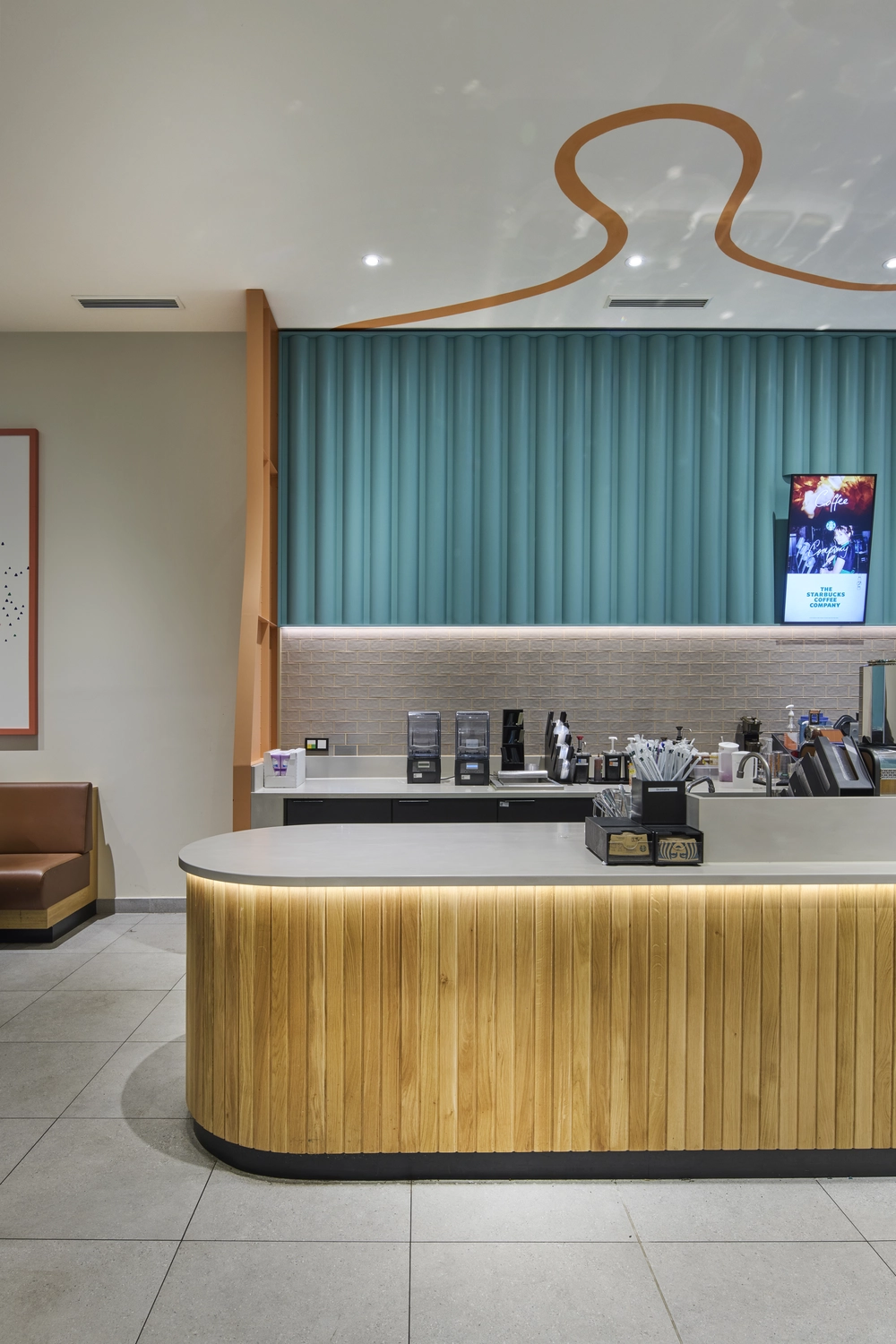 Heinrich Buchholz – Holzbearbeitung – Moderne Café-Theke mit geschwungener Front aus Naturholzlamellen und warmer LED-Unterbaubeleuchtung vor einer türkisfarbenen, geriffelten Akzentwand.