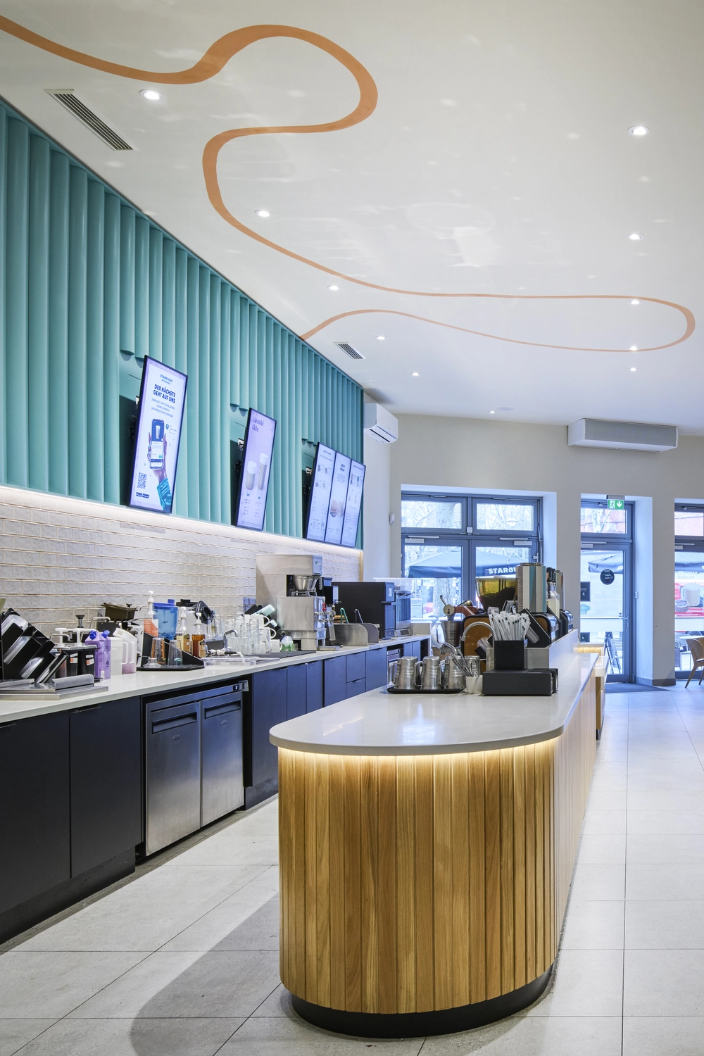 Heinrich Buchholz – Holzbearbeitung – Modernes Café-Interieur mit einer langen, geschwungenen Theke, deren Fassade aus Naturholzlamellen besteht und die über eine warme LED-Unterbauleuchte verfügt. Dazu passend ein hölzerner Produktpräsentationsständer.