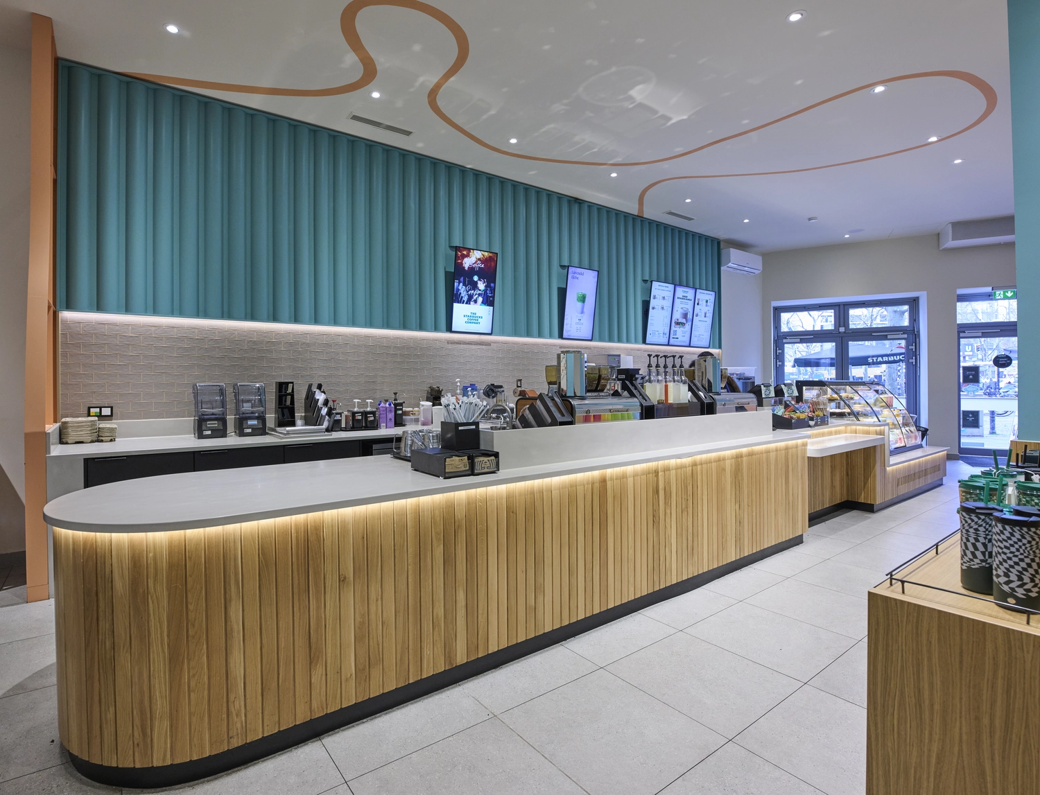 Heinrich Buchholz – Holzbearbeitung – Modernes Café-Interieur mit einer langen, geschwungenen Theke, deren Fassade aus Naturholzlamellen besteht und die über eine warme LED-Unterbauleuchte verfügt. Dazu passend ein hölzerner Produktpräsentationsständer.