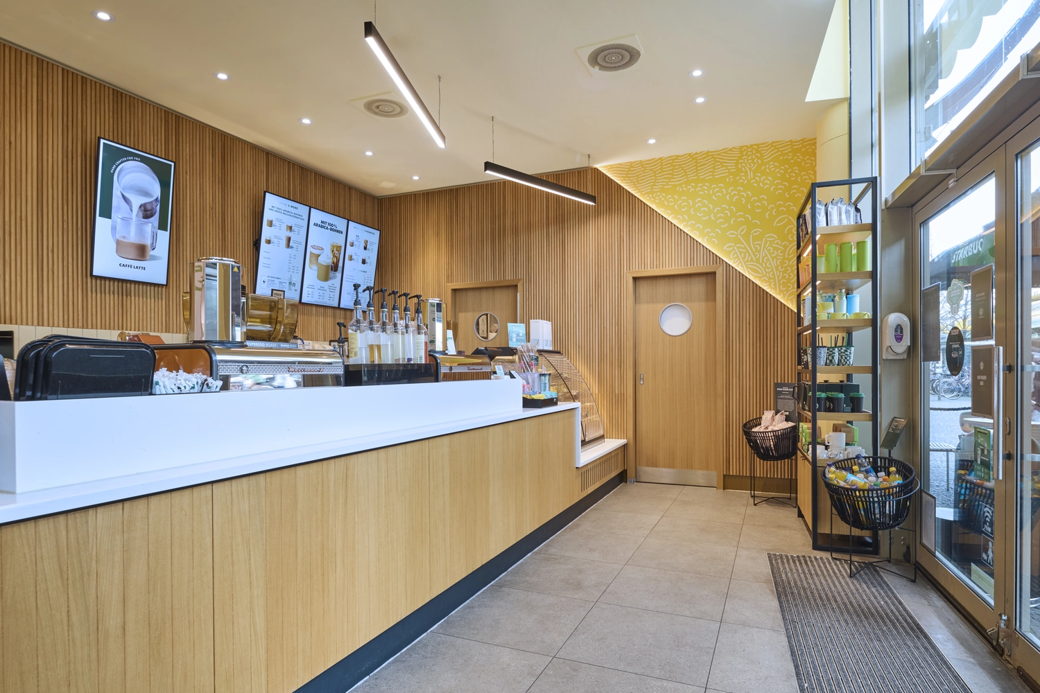 Heinrich Buchholz – Holzbearbeitung – Modernes Café-Interieur mit einer langen Holztheke mit weißer Arbeitsplatte, passender Wandverkleidung aus Eichenholzlamellen und maßgefertigten Türen.