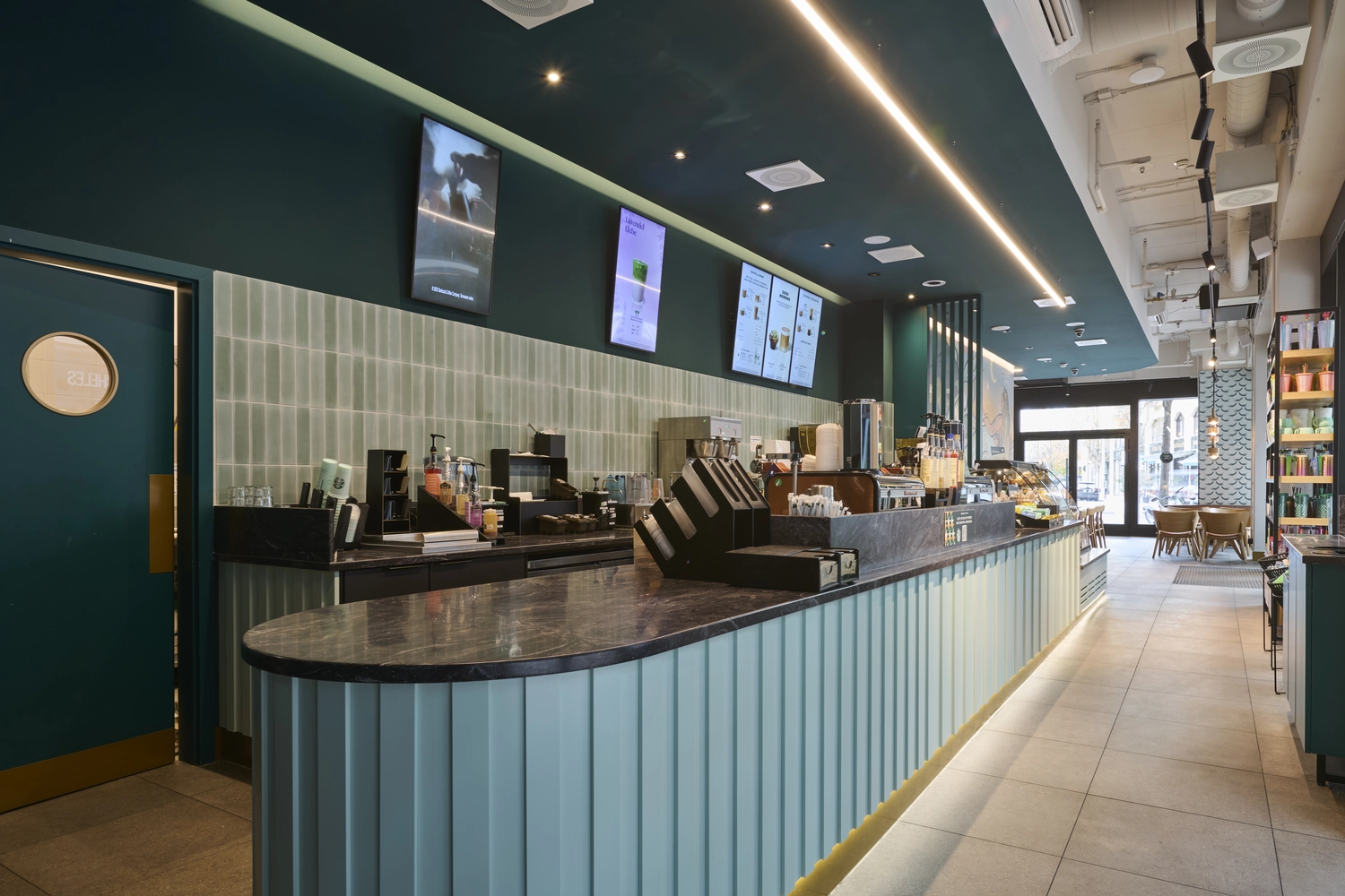 Heinrich Buchholz – Holzbearbeitung – Modernes Café-Interieur mit einer langen, türkisfarbenen Theke, deren geschwungene, geriffelte Paneelfassade, eine dunkle Marmorarbeitsplatte und eine dazu passende, maßgefertigte türkisfarbene Tür mit rundem Bullauge verfügen.