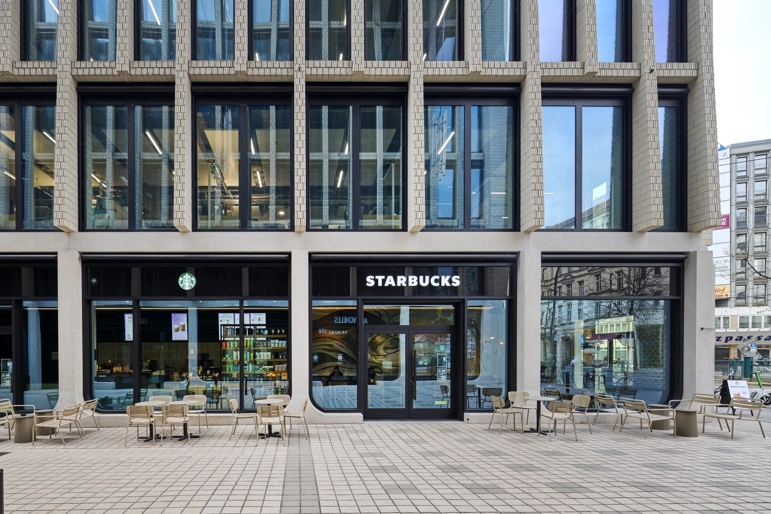 Heinrich Buchholz – Holzbearbeitung – Moderne Ladenfront eines Geschäftsgebäudes mit einem Starbucks-Café, großen Glasfronten, elegantem schwarzem Holzrahmen und gepflastertem Außenbereich.